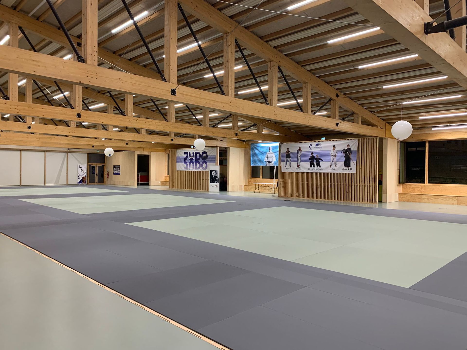 Intérieur du nouveau dojo du Judo Club de Hem : grande salle à charpente en bois, tatamis gris et vert clair, éclairage LED intégré à la charpente, avec en fond les affiches des disciplines proposées par le club (judo, ju-jitsu, taïso, kendo).